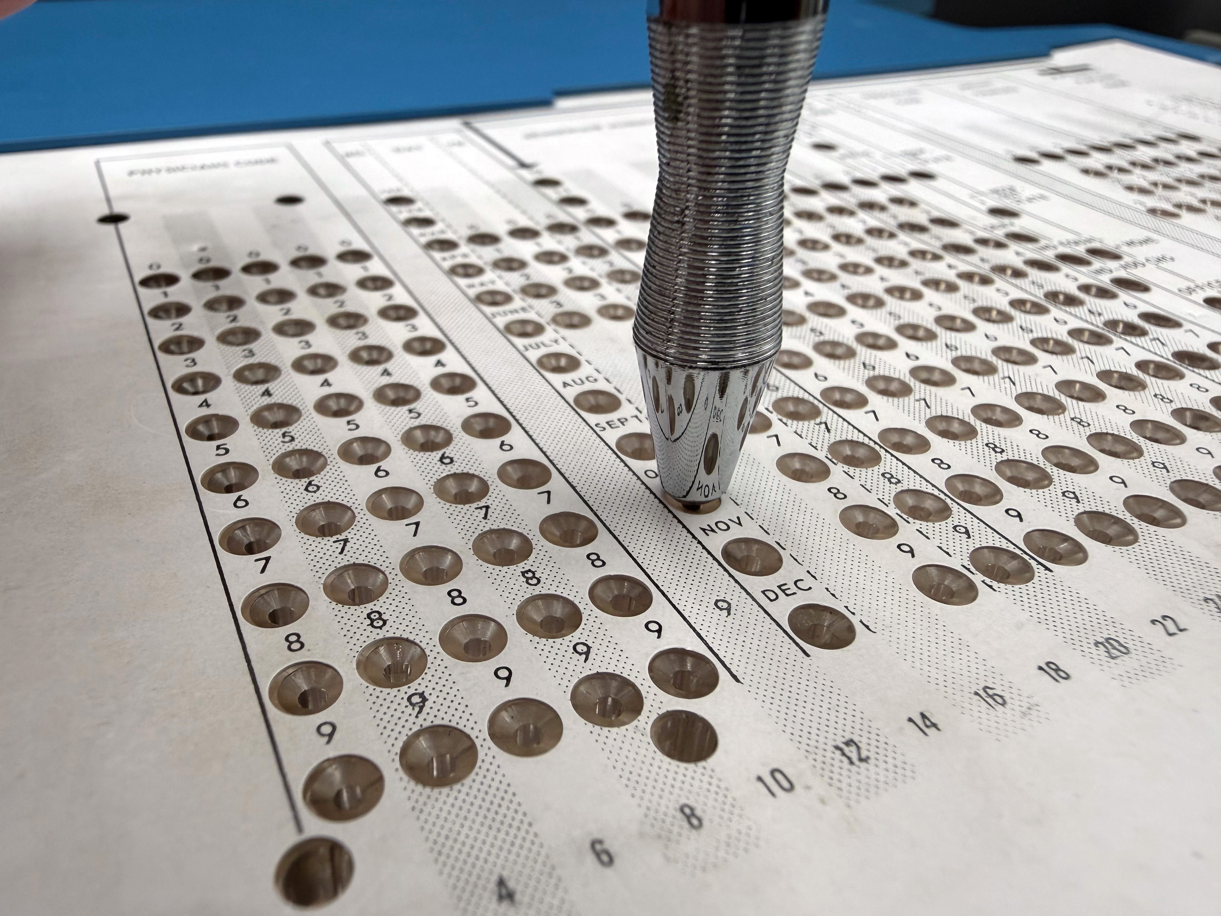 Detail showing the stylus inserted in the punch, punching the "October" position of the Month field on the template overlay.